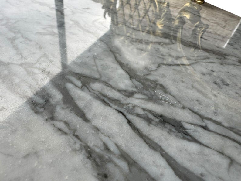 May include: Close-up of a white marble surface with gray veining. The polished stone reflects light, creating a glossy appearance. The surface is part of a larger piece, possibly a table or countertop.