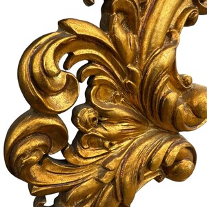 May include: Ornate, gold-colored decorative architectural element with a detailed, swirling leaf and scroll design. The piece has a distressed, antique appearance, with visible texture and variations in the gold finish.