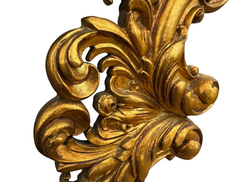 May include: Ornate, gold-colored decorative architectural element with a detailed, swirling leaf and scroll design. The piece has a distressed, antique appearance, with visible texture and variations in the gold finish.