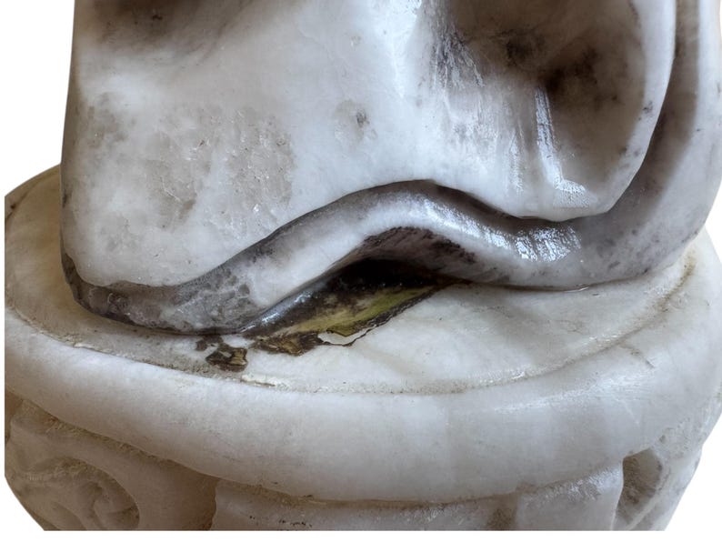 K&ouml;nnte beinhalten: Nahaufnahme eines wei&szlig;en Marmor-Skulpturenfragments. Die Skulptur hat eine geschwungene, glatte Form mit dunklem Akzent. Der Sockel ist rund mit geschnitzten Details.
