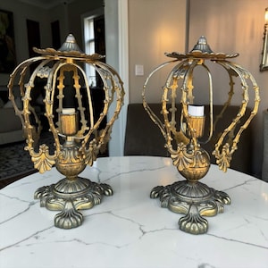 May include: A pair of gold-toned metal table lamps featuring an open, cage-like crown structure with ornate, curved ribs and a pointed finial top. Each lamp has a central light bulb socket and a decorative, wide base with four feet.