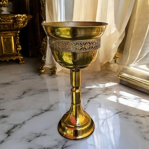 Puede incluir: Un cáliz de latón dorado con un cuenco ancho y una banda decorativa con un intrincado diseño. El cáliz se apoya en un tallo delgado y una base acampanada. La superficie pulida refleja la luz circundante, creando un brillo cálido.