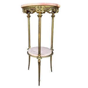 May include: An antique plant stand with a marble top and a lower shelf. The stand has a gold-coloured metal frame with ornate floral detailing. The top is oval-shaped, and the stand has three legs.