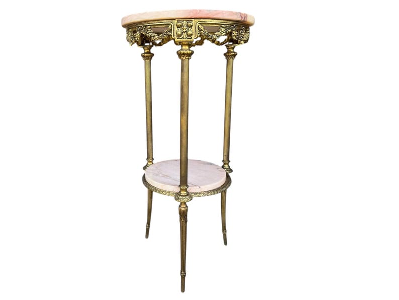 May include: An antique plant stand with a marble top and a lower shelf. The stand has a gold-coloured metal frame with ornate floral detailing. The top is oval-shaped, and the stand has three legs.