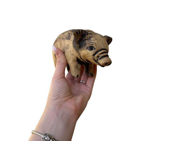 Pu&ograve; includere: Una piccola statuetta di maiale in ceramica beige con dettagli marrone scuro. Il maiale ha tratti dettagliati, tra cui orecchie, muso e occhi visibili. La statuetta &egrave; tenuta su uno sfondo bianco.
