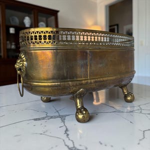 Puede incluir: Una jardinera antigua de latón con un borde calado decorativo y asas con cabeza de león. La jardinera de forma ovalada descansa sobre tres patas de bola y garra. El latón tiene una pátina cálida y envejecida. Una pieza clásica para la decoración del hogar.