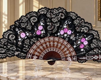 Op de afbeelding: Een vouwwaaier met een donkerbruin houten frame met ingewikkelde witte filigraanpatronen en een zwarte kanten waaierblad. Het kant is versierd met geborduurde roze bloemtrossen en kleine witte bloesems. Het nummer 114854 staat op het handvat gedrukt.