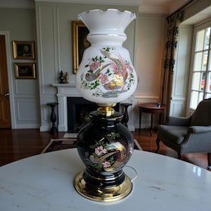 Puede incluir: Una lámpara de mesa vintage con base negra y pantalla de cristal blanca. La lámpara presenta una base y detalles dorados, y está decorada con coloridos diseños de pájaros y flores. Es una lámpara decorativa.