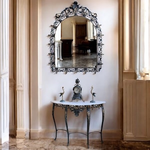 May include: An ornate, arched mirror with a dark metal frame hangs above a marble-topped console table. The table features a decorative metal base. Two bird figurines and a clock sit on the table. The room has a classic, elegant style.