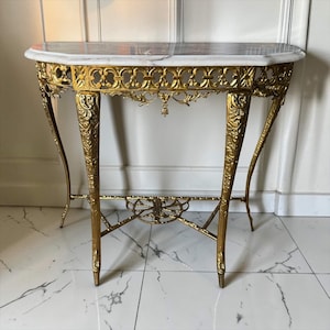 May include: A semi-circular console table featuring a white marble top with grey veining. The frame is constructed from gold-toned metal with intricate filigree detailing along the apron and four ornate, tapered legs connected by a central stretcher.