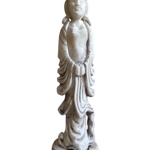 K&ouml;nnte beinhalten: Verwitterte, cremefarbene Statue einer friedlichen Figur in flie&szlig;enden Gew&auml;ndern. Die Statue weist aufwendige Details auf, darunter eine Kopfbedeckung und ein verzierter Sockel. Die H&auml;nde der Figur sind vor ihr gefaltet.