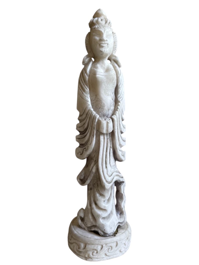 K&ouml;nnte beinhalten: Verwitterte, cremefarbene Statue einer friedlichen Figur in flie&szlig;enden Gew&auml;ndern. Die Statue weist aufwendige Details auf, darunter eine Kopfbedeckung und ein verzierter Sockel. Die H&auml;nde der Figur sind vor ihr gefaltet.