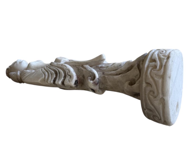 K&ouml;nnte beinhalten: Verwitterte, cremefarbene Statue einer klassischen Figur, m&ouml;glicherweise einer G&ouml;ttin oder eines Engels, mit detaillierten Fl&uuml;geln und drapierter Kleidung. Die Statue hat einen rechteckigen Sockel mit kunstvollen Schnitzereien.