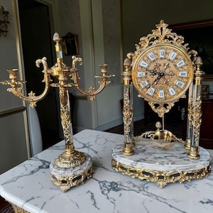 May include: A gold-toned metal candelabra and a matching mantel clock, both featuring clear acrylic columns and white marble bases with ornate gold filigree detailing. The clock has a round face with black Roman numerals and a gold pendulum.