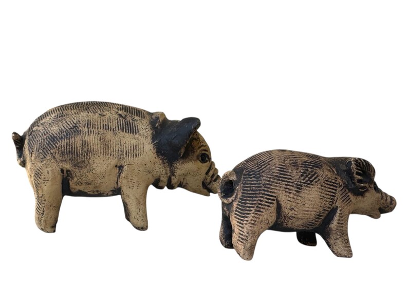Pu&ograve; includere: Due statuette di maiali in ceramica. I maiali sono color beige con accenti neri sulle orecchie e sui musi. Le statuette hanno dettagli strutturati e sono posizionate come se interagissero tra loro. Lo sfondo &egrave; bianco.