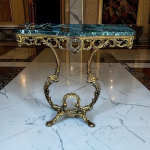 May include: A console table featuring a rectangular dark green marble top with white veining. The base is constructed from ornate gold-toned metal with intricate scrollwork, curved legs, and a central decorative medallion element.