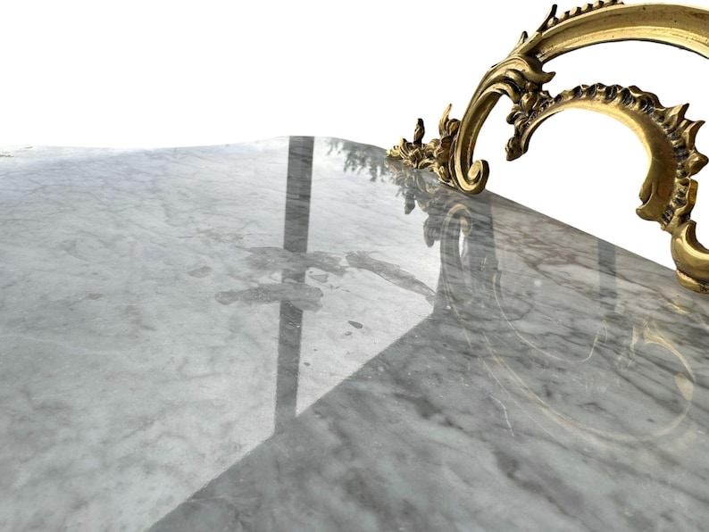 May include: Close-up of a marble surface with gray and white veining. A decorative gold-colored metal accent with ornate scrollwork is visible in the upper right corner. The surface reflects light.