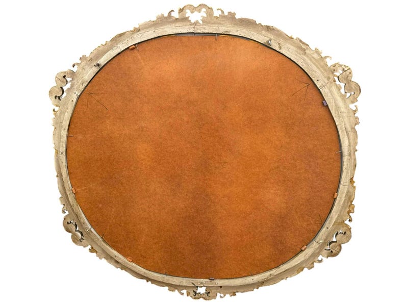 May include: An antique, oval-shaped mirror with a gold-colored, ornate frame. The back of the mirror is visible, featuring a warm, brown backing. The frame has detailed scrollwork and decorative elements.