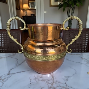 Puede incluir: Un jarrón de cobre con dos asas doradas ornamentadas. El jarrón tiene una textura martillada y una banda dorada decorativa en el medio. El jarrón está sobre una superficie de mármol blanco.