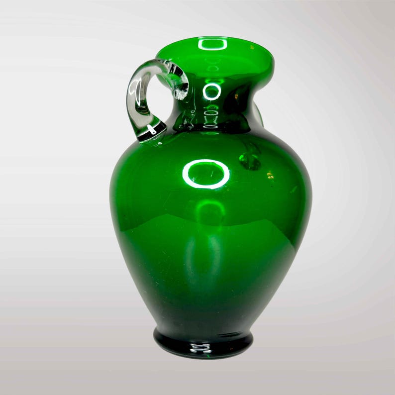 May include: A dark green glass vase with a clear glass handle. The vase has a rounded body and a narrow neck.