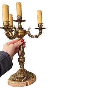Pu&ograve; includere: Candelabro in ottone ornato con quattro luci a finto candela. Il candelabro ha un design vintage dettagliato con una base in marmo. La mano che tiene il candelabro indossa una giacca scura.