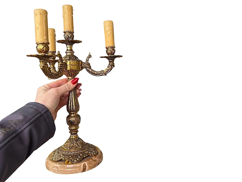 Pu&ograve; includere: Candelabro in ottone ornato con quattro luci a finto candela. Il candelabro ha un design vintage dettagliato con una base in marmo. La mano che tiene il candelabro indossa una giacca scura.