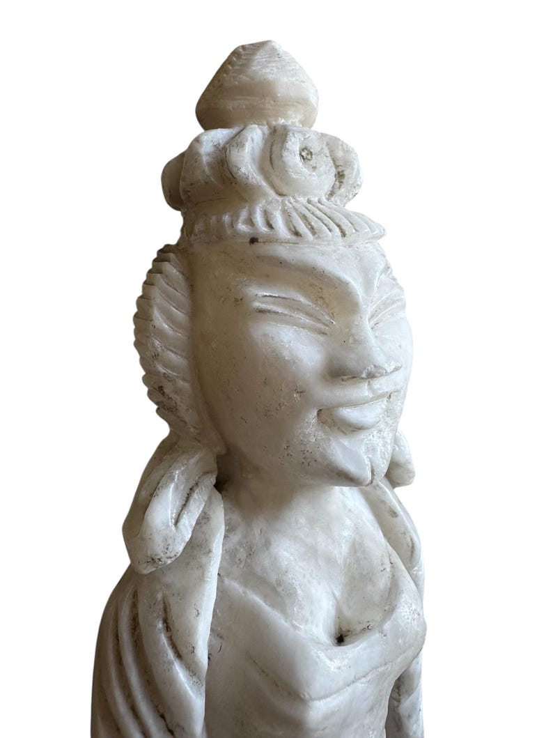K&ouml;nnte beinhalten: Eine geschnitzte Statue aus wei&szlig;em Marmor einer ruhigen Figur mit aufwendigem Kopfschmuck und flie&szlig;enden Gew&auml;ndern. Die Statue hat ein sanftes L&auml;cheln und detaillierte Gesichtsz&uuml;ge, die die Handwerkskunst der Skulptur zeigen.