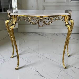Può includere: Un tavolo console caratterizzato da un piano in marmo bianco liscio con finitura lucida. La base è realizzata in metallo dorato decorato con intricati motivi a volute e quattro gambe sottili e curve che si restringono verso il pavimento.