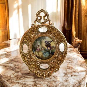 May include: Ornate gold-colored decorative plate with a central painted scene of figures in a garden. The plate has a circular shape with decorative scrollwork and a handle at the top. The painting depicts a man playing a lute for two women.