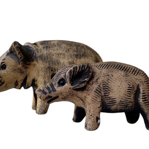 Pu&ograve; includere: Due statuette di cinghiali in ceramica. Il cinghiale pi&ugrave; grande &egrave; dietro quello pi&ugrave; piccolo. Entrambi sono beige con dettagli marrone scuro, tra cui occhi, musi e linee incise sui loro corpi. Su sfondo bianco.