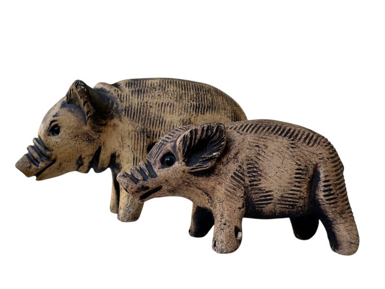 Pu&ograve; includere: Due statuette di cinghiali in ceramica. Il cinghiale pi&ugrave; grande &egrave; dietro quello pi&ugrave; piccolo. Entrambi sono beige con dettagli marrone scuro, tra cui occhi, musi e linee incise sui loro corpi. Su sfondo bianco.
