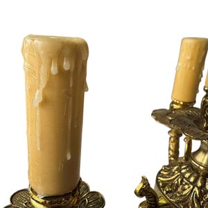 Pu&ograve; includere: Primo piano di un candelabro vintage in ottone con tre portalampada. I candelabri hanno dettagli ornati e contengono candele color crema con gocce di cera. Lo sfondo &egrave; bianco.