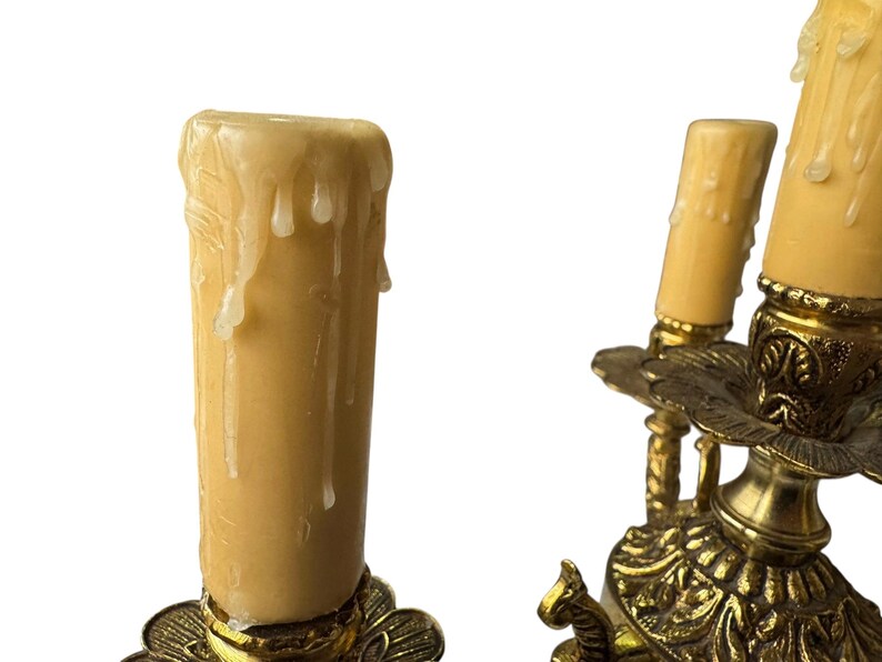 Pu&ograve; includere: Primo piano di un candelabro vintage in ottone con tre portalampada. I candelabri hanno dettagli ornati e contengono candele color crema con gocce di cera. Lo sfondo &egrave; bianco.
