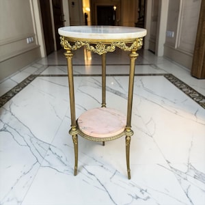 May include: An antique, two-tiered side table with a marble top and a lower shelf. The table has a gold-coloured metal frame with ornate floral detailing. The top shelf is white marble, and the lower shelf is pink marble.