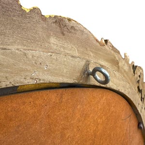 May include: Close-up of a decorative, ornate mirror frame. The frame is a light, neutral color with gold accents and a textured surface. A metal ring is attached for hanging. The mirror's backing is a warm, reddish-brown color.