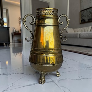 Op de afbeelding: Een vintage messing urn met sierlijke handvatten en een decoratief rooster. De urn heeft een bolvormig lichaam en staat op drie klauwpoten. Het gepolijste messing reflecteert licht, waardoor een warme, gouden gloed ontstaat. Geschikt voor interieurdecoratie.