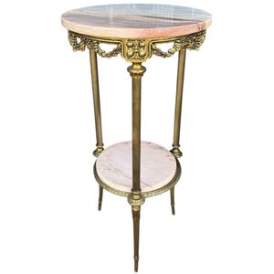 May include: A vintage plant stand with a round, pink and white marble top and a lower shelf. The stand features a gold-coloured metal frame with ornate detailing and three slender legs. The stand is approximately 76 cm tall.