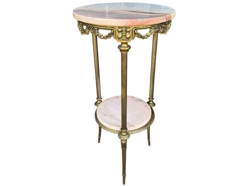 May include: A vintage plant stand with a round, pink and white marble top and a lower shelf. The stand features a gold-coloured metal frame with ornate detailing and three slender legs. The stand is approximately 76 cm tall.