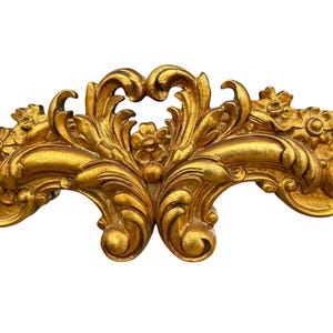 May include: Ornate gold-colored decorative architectural element with a symmetrical design. The element features floral and scroll motifs, with a detailed, textured surface. The piece is likely part of a larger decorative frame or molding.