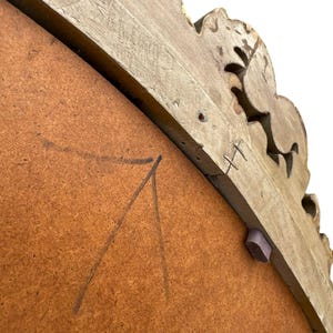 May include: Close-up of the back of a decorative mirror. The frame is light brown with carved floral details. The mirror backing is a medium brown color with pencil markings. A small, purple wooden piece is attached to the frame.