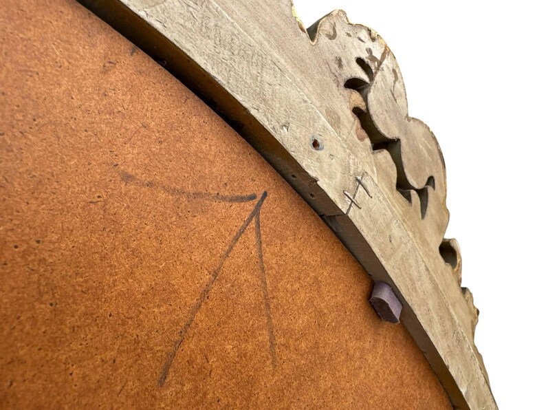 May include: Close-up of the back of a decorative mirror. The frame is light brown with carved floral details. The mirror backing is a medium brown color with pencil markings. A small, purple wooden piece is attached to the frame.