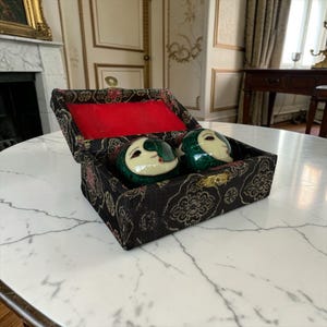 Peut inclure: Une boîte noire avec un intérieur rouge contient deux sphères décoratives vertes et blanches avec des visages. La boîte a un fermoir doré et un motif floral. Les sphères présentent un motif de croissant de lune.