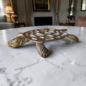 Puede incluir: Un salvamanteles de latón con forma de tortuga que presenta cabeza y patas texturizadas junto a un diseño de caparazón calado detallado. El objeto de metal tiene un acabado pulido y tiene la forma de una tortuga marina con cuatro patas.