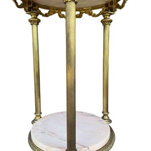 May include: An ornate side table in gold tones, featuring a marble top and base. The table has three fluted legs and decorative trim. The marble displays light pink and white veining. The table's height is approximately 76 cm.