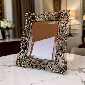 May include: An antique-style picture frame with a bronze finish. The frame has elaborate scrollwork and a rectangular opening with a glass cover. The frame is displayed on a white marble surface.