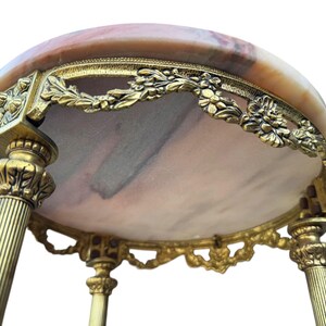May include: An antique side table with a round marble top in peach and brown hues. The table has ornate, gold-coloured metalwork around the edge and on the legs, with floral and leaf designs. The table has three legs.