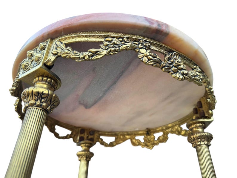 May include: An antique side table with a round marble top in peach and brown hues. The table has ornate, gold-coloured metalwork around the edge and on the legs, with floral and leaf designs. The table has three legs.