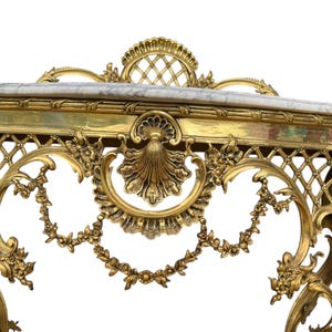 May include: Ornate gold-colored console table with a marble top. The table features intricate floral and scrollwork detailing, including a lattice design and a shell-shaped central motif. The overall design is classical and elegant.