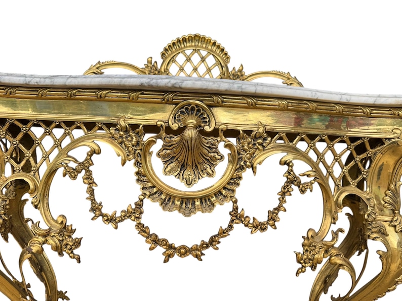 May include: Ornate gold-colored console table with a marble top. The table features intricate floral and scrollwork detailing, including a lattice design and a shell-shaped central motif. The overall design is classical and elegant.