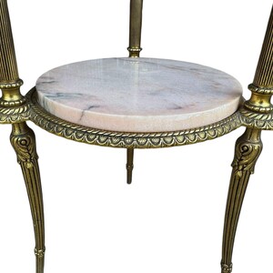 May include: An antique side table with a round, pink marble top. The table has a brass-coloured metal frame with ornate detailing and three curved legs. The table is designed to hold decorative items.